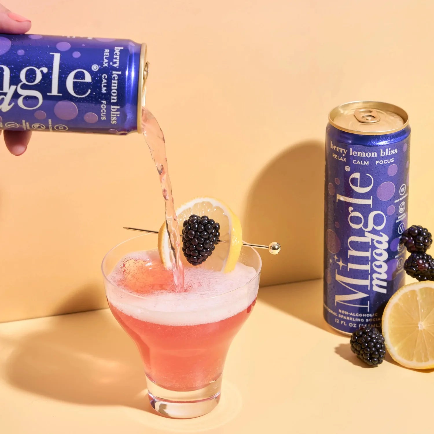 Hand pouring Mingle Mood Berry Lemon Bliss from a navy blue can into a cocktail glass with blackberry and lemon garnish.