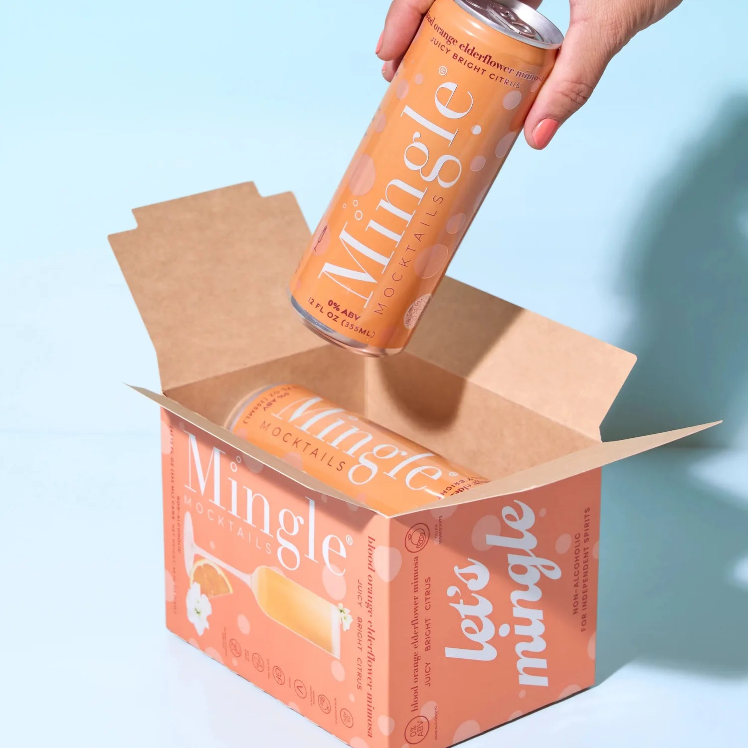 Hand pulling a Mingle Mocktails Blood Orange Elderflower Mimosa can from a branded peach shipping box on a light blue background.