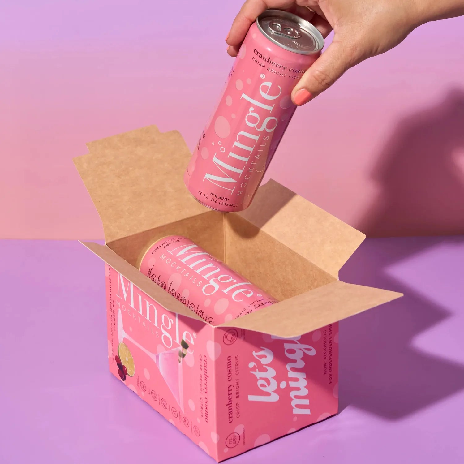 Hand pulling a Mingle Mocktails Cranberry Cosmo can from a branded pink shipping box on a purple background.
