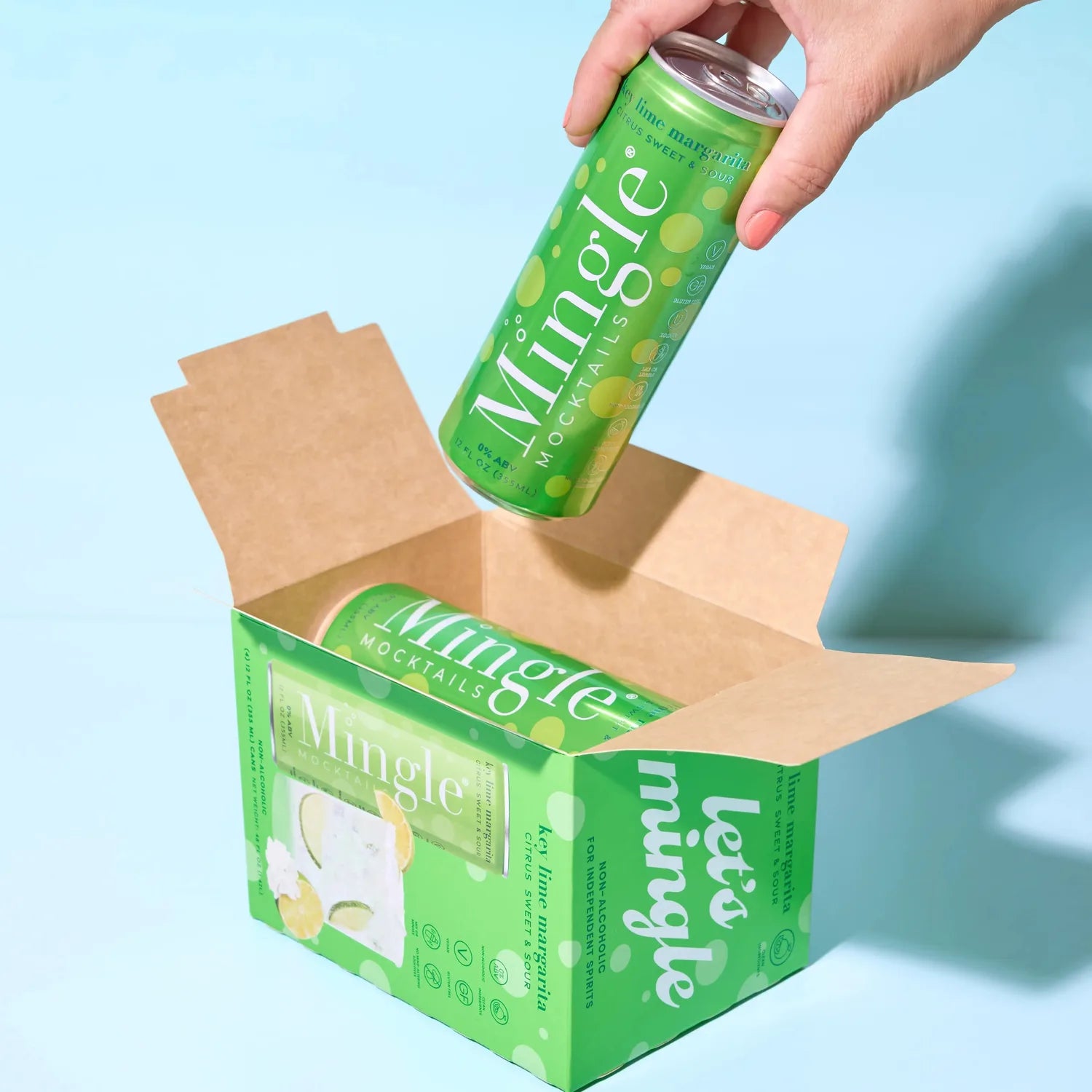Hand pulling a Mingle Mocktails Key Lime Margarita can from a branded green shipping box on a light blue background.