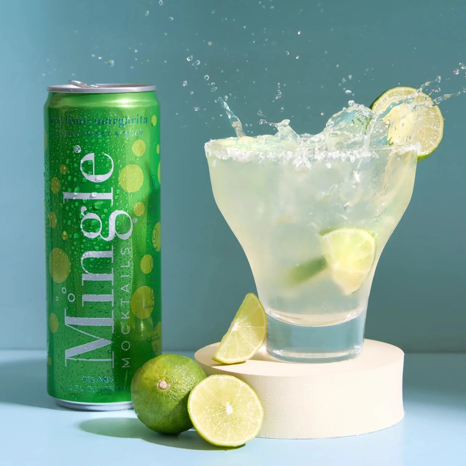 Mingle Mocktails Key Lime Margarita green can beside a splashing margarita glass with lime wheels on a teal background.
